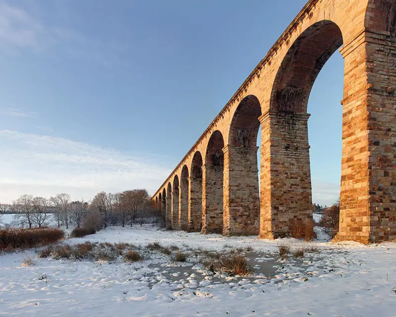 Sunlight illuminates the Crimple Viaduct in winter . a snow covered bridge in front of a brick building. sky outdoor building bridge snow brick cloud arch winter