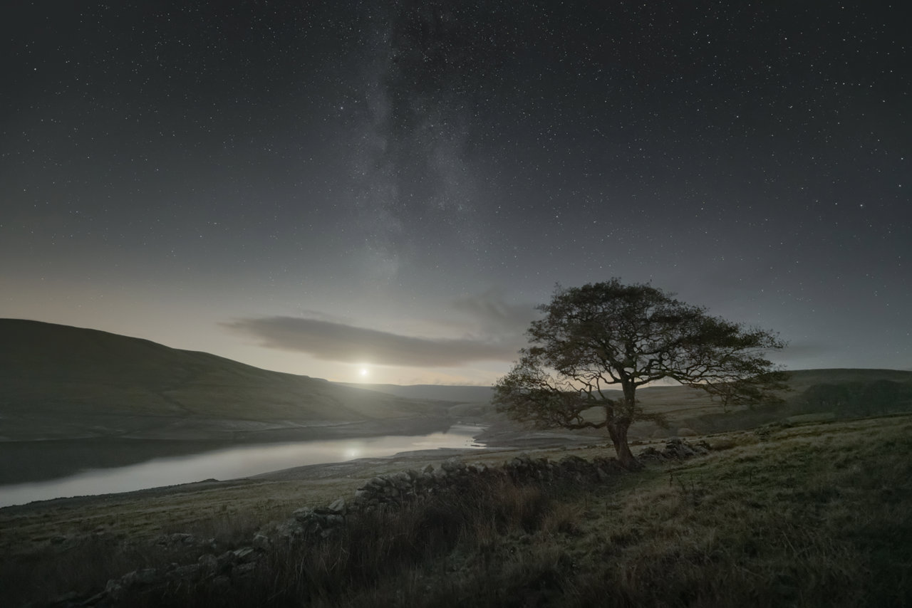 A solitary tree stands in the serene countryside of North Yorkshire at night. Rolling hills frame a tranquil reservoir, its surface reflecting the light of a near-full moon nestled on the horizon. Above, a clear sky reveals the Milky Way's cosmic splendour, stars dotting the expanse. A stone wall gently meanders across the grassy foreground.