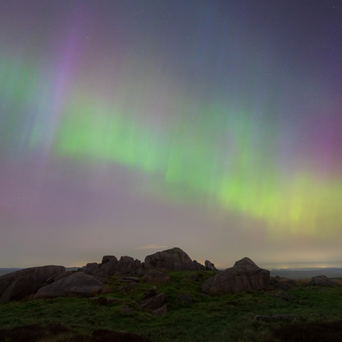 Aurora Borealis above moorland outcrop: North Yorkshire moorland landscape under the Northern Lights. Elegant drapes of green and purple hues dance across the heavens, casting a surreal glow over the earthy terrain scattered with rocks and unassuming grasses below.
