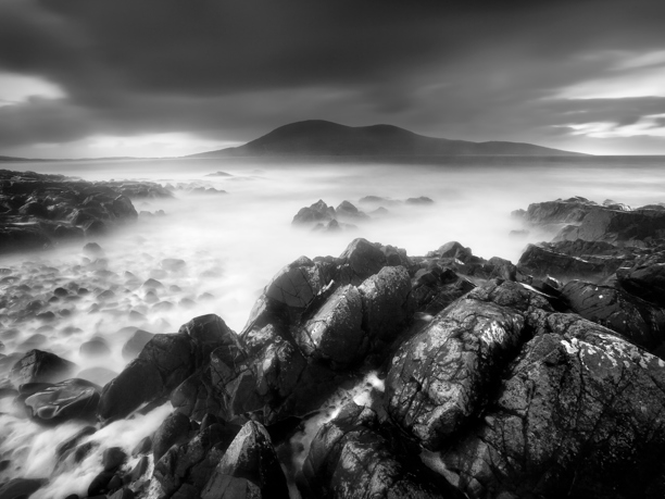 Fluid A monochrome seascape depicts a smooth mist over the water, with jagged rocks in the foreground. In the distance, a mountain looms beneath brooding clouds, creating a serene and dramatic atmosphere.