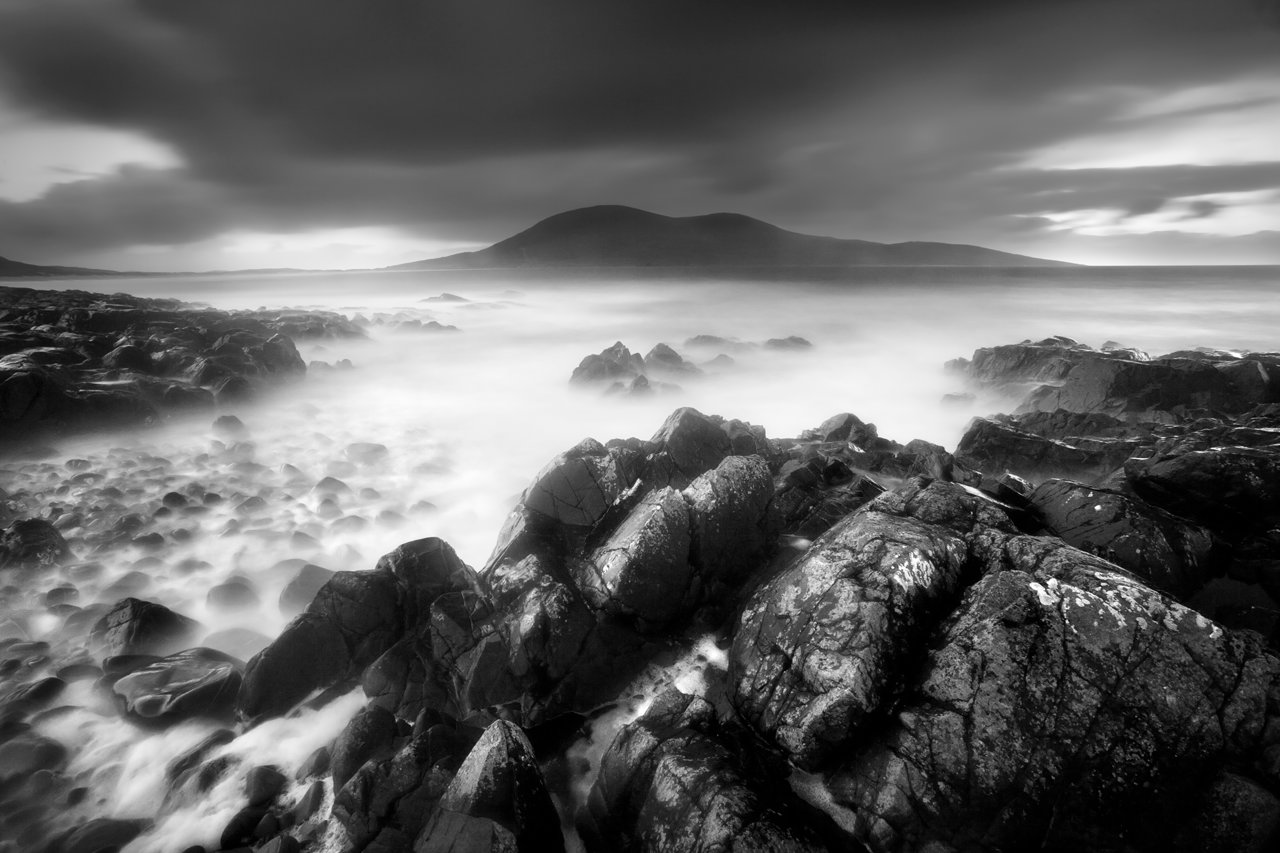 A monochrome seascape depicts a smooth mist over the water, with jagged rocks in the foreground. In the distance, a mountain looms beneath brooding clouds, creating a serene and dramatic atmosphere.
