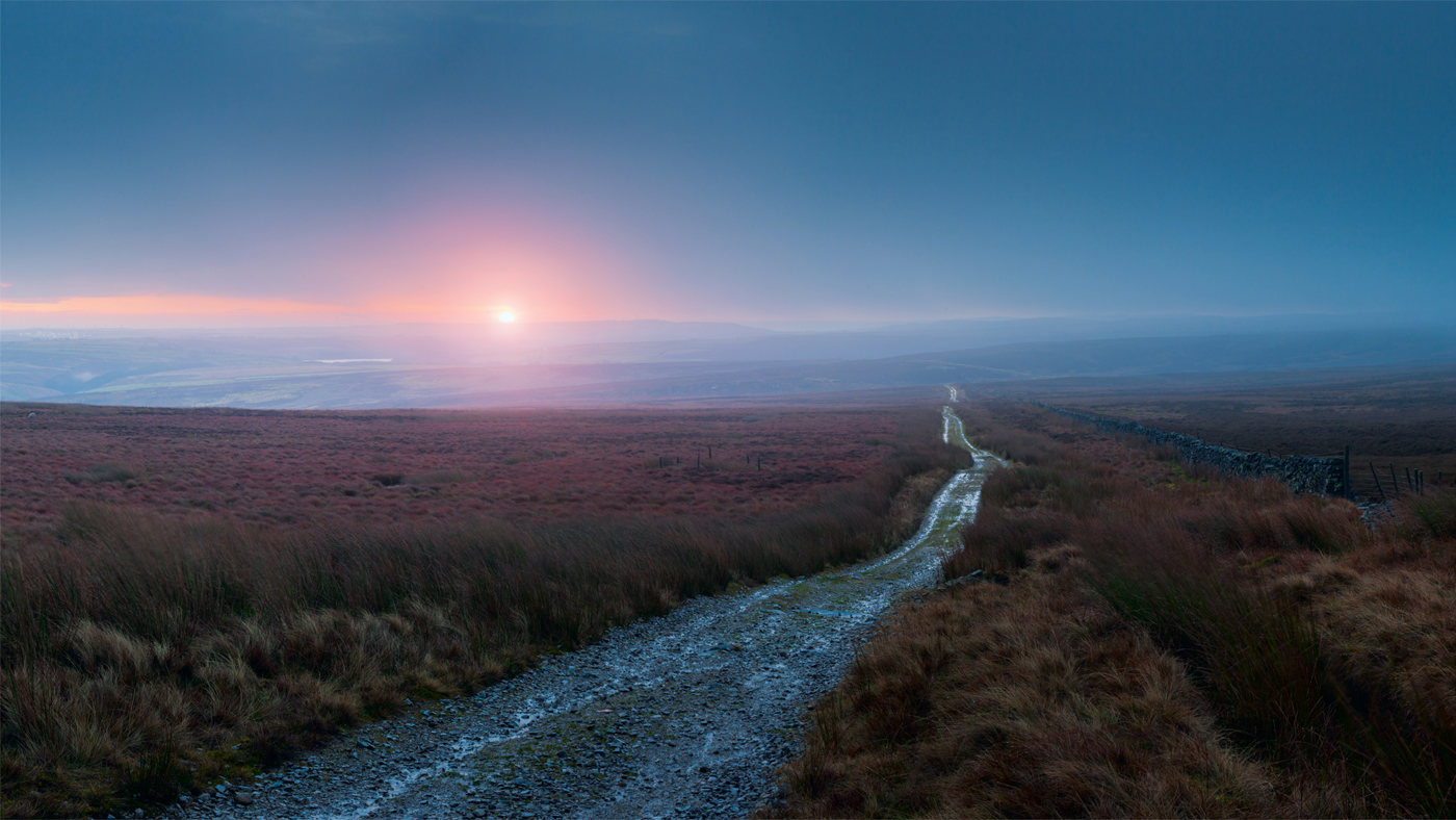 The image shows a serene North Yorkshire moor at dawn. A gravel path meanders through the heather-filled terrain, leading toward the horizon under a pastel sky. The rising sun casts a warm glow. The moor appears tranquil and expansive.