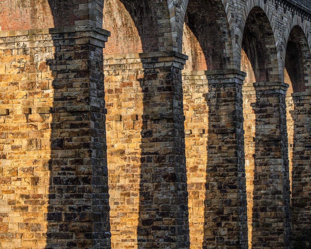 The Crimple Valley Viaduct's stone structure bathed in sunlight. Arches rise from sturdy pillars, their shadows striping the textured walls. The golden hue of the stone contrasts with the soft arch shadows, evoking a sense of historic grandeur. a large brick building