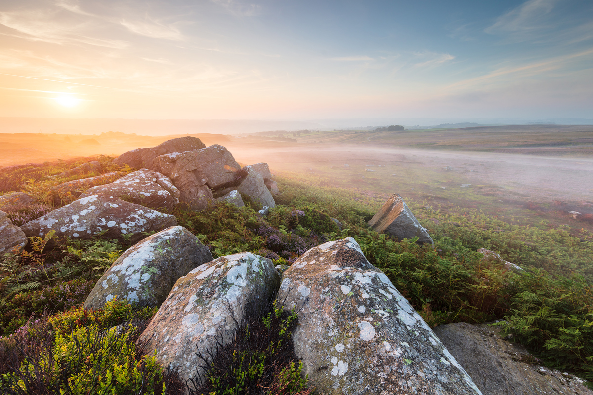 An intense sunrise bathes North Yorkshire moorland in soft light. Lichen-speckled rocks and heather streak the foreground, while a misty expanse stretches towards a hazy horizon. a rocky landscape with a valley in the background