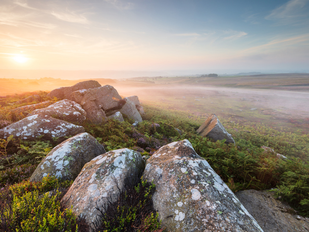 An intense sunrise bathes North Yorkshire moorland in soft light. Lichen-speckled rocks and heather streak the foreground, while a misty expanse stretches towards a hazy horizon.
