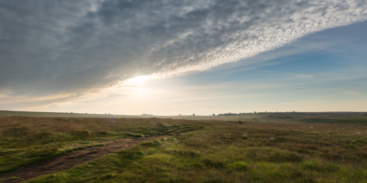 You're looking at a serene North Yorkshire moor at dusk. A worn path trails through the undisturbed grassland leading towards the horizon. The sky is a dramatic display with the sun partially veiled by wisps of cloud, casting a soft golden glow over the tranquil scene. a grassy field with clouds in the sky