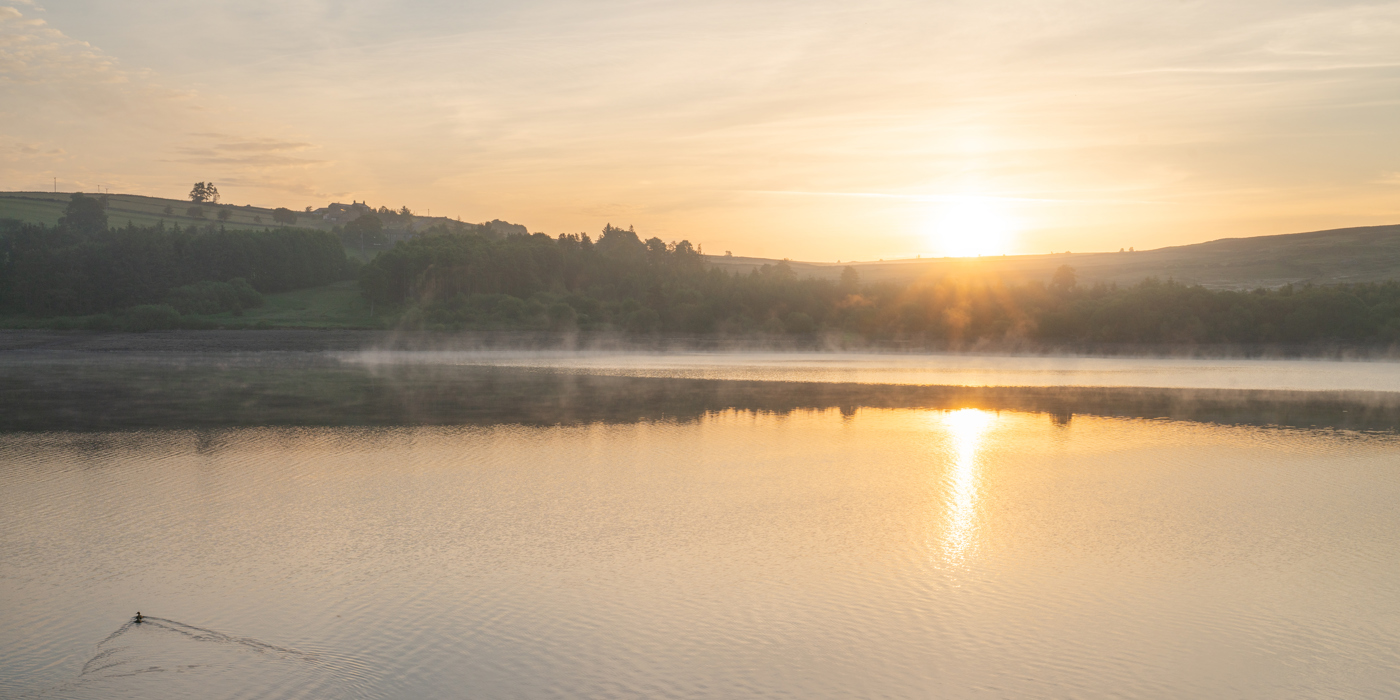 A serene North Yorkshire sunrise reflects on a calm lake. Mist hovers above the water's surface. The sun casts a warm glow, silhouetting hills and trees against a soft sky. A lone waterbird trails ripples across the golden stillness. a sunset over a body of water