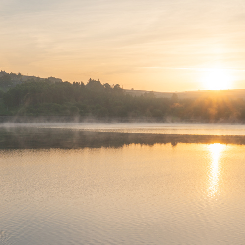 15th June 2022: A serene North Yorkshire sunrise reflects on a calm lake. Mist hovers above the water's surface. The sun casts a warm glow, silhouetting hills and trees against a soft sky. A lone waterbird trails ripples across the golden stillness.