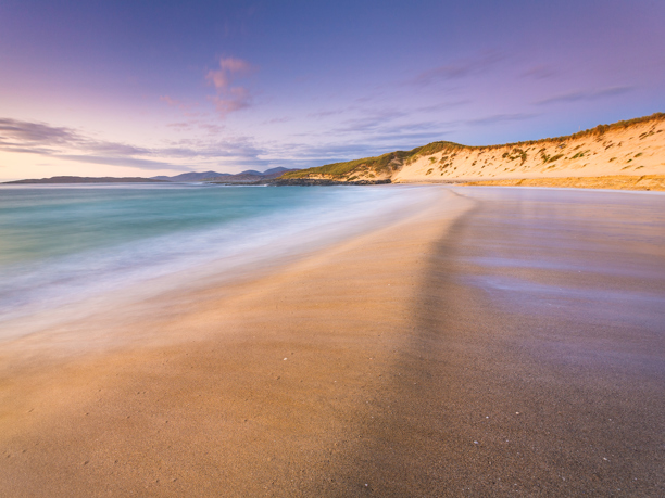 A sweeping sandy beach graces the foreground, with gentle waves caressing the shore and leaving a smooth, wet surface. A grass-topped dune rises to the right, bathed in warm light from a softly lit sky streaked with lavender and gold hues. In the distance, gentle hills fade into the horizon under a tranquil sky.