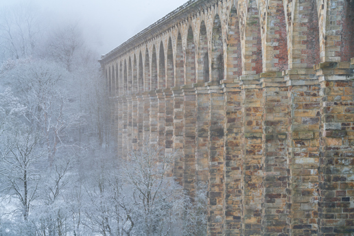 The Crimple Viaduct cloaked in winter's embrace. It stands majestically with arches in a wintry North Yorkshire forest. The trees, dusted in snow, emerge from a blanket of fog, enhancing the viaduct's old-world charm. a brick wall with snow on it